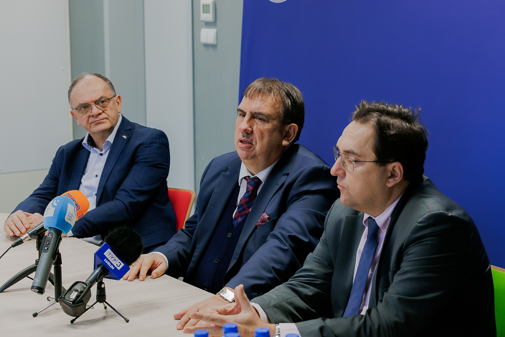 Trzech mężczyzn w garniturach siedzących za stołem, na którym są mikrofony dziennikarskie