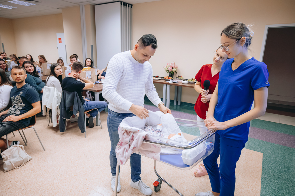 Po lewej stronie, przy noworodkowym łóżeczku szpitalnym stoją: jeden z uczestników spotkania – mężczyzna jest pochylony nad lalką imitującą dziecko oraz prowadzące warsztaty z podstaw pielęgnacji Paulina Ciorgoń i Dominika Rafał. Z prawej strony siedzący przy stołach uczestnicy spotkania. 