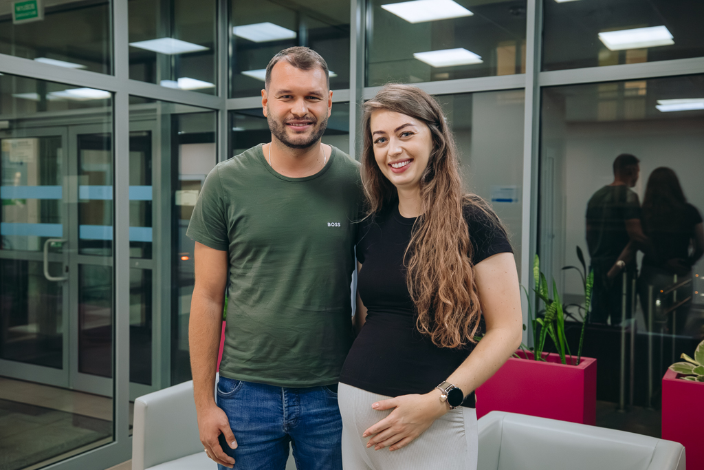Uczestnicy spotkania - stojący obok siebie pani Aleksandra i Pan Piotr, na tle okna przy wejściu do budynku Bloku Operacyjnego. W tle dwa szare fotele i wysokie doniczki z kwiatkami. 