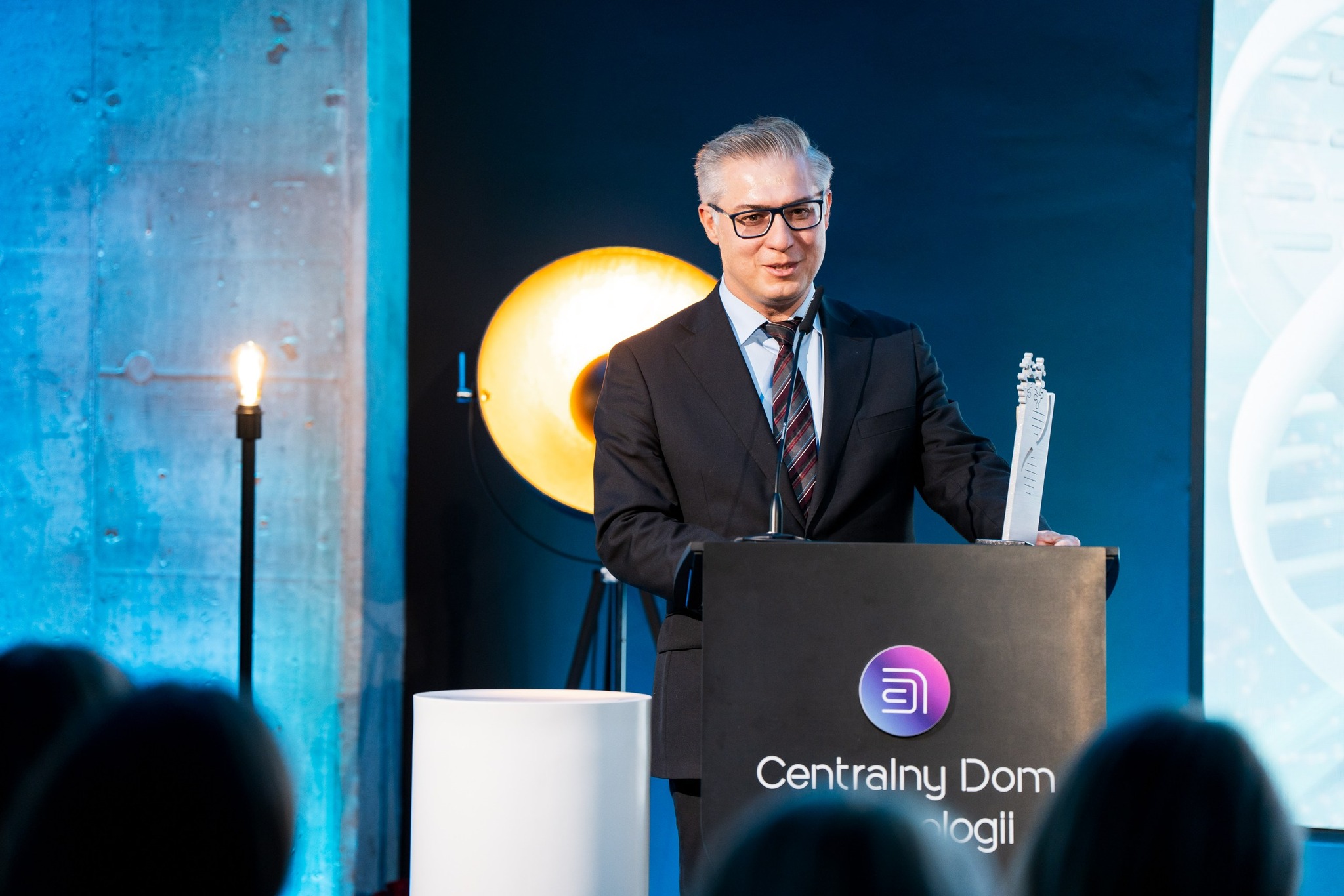 Na fotografii widać mężczyznę stojącego za mównicą podczas wystąpienia. Ma na sobie ciemny garnitur, koszulę i krawat oraz okulary. Jedną ręką opiera się o mównicę, a w drugiej trzyma smukłą statuetkę. Na przodzie mównicy znajduje się znak i niewyraźny napis Centralny Dom Technologii. W tle widać ciemnoniebieską ścianę i dużą okrągłą lampę świecącą ciepłym światłem. Po lewej stronie mężczyzny na statywie stoi zapalona żarówka.  