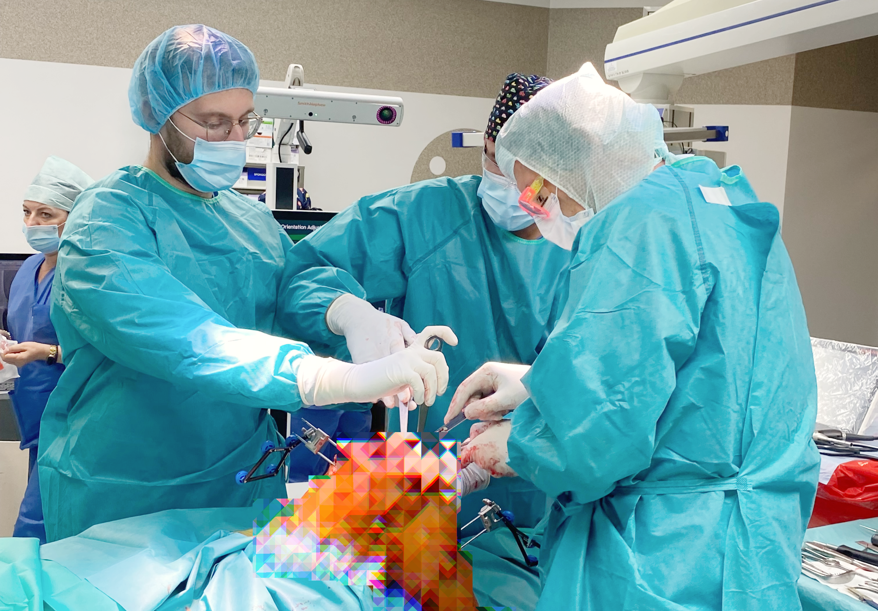 Trzech medyków przy pomocy narzędzi operuje na sali operacyjnej kolano chorego, które jest zamazane z powodu drastyczności obrazu. Z tyłu jeszcze jeden medyk. Wszyscy w pełnej odzieży medycznej.