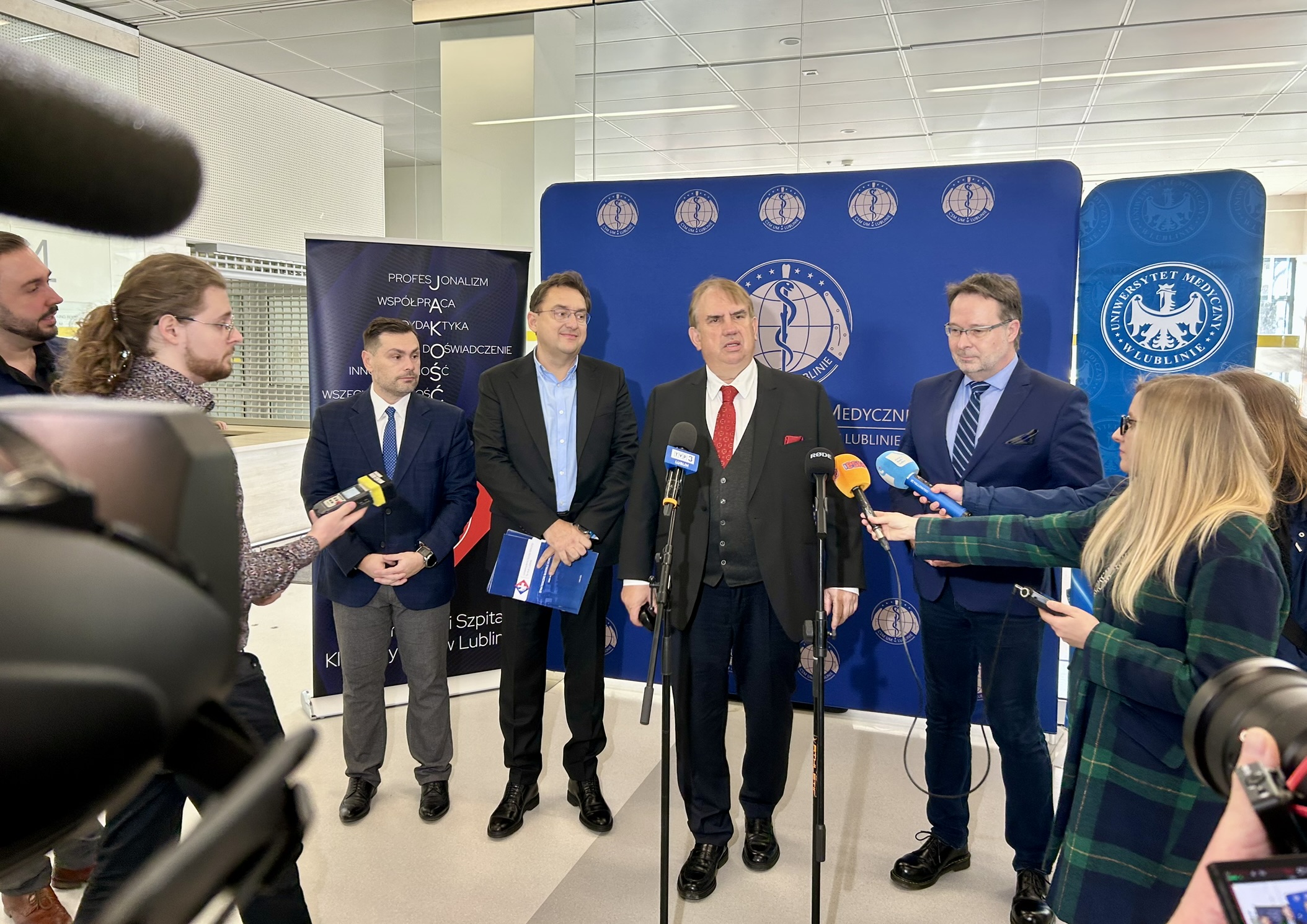 Po lewej stronie fotografii widać fragment sprzętu do nagrywania oraz kilka osób stojących bokiem i trzymających w rękach mikrofony skierowane w stronę grupy ludzi stojących na środku. Na środku zdjęcia stoi pięciu mężczyzn ustawionych w jednym rzędzie. Wszyscy mają na sobie garnitury lub marynarki i patrzą w stronę osób z mikrofonami. Mężczyzna stojący pośrodku ma ciemny garnitur, białą koszulę i czerwony krawat, a przed nim ustawione są dwa stojaki z mikrofonami. Obok niego po lewej i prawej stronie stoją pozostali mężczyźni, jeden z nich trzyma w ręku niebieską teczkę. Za nimi znajduje się duża niebieska ścianka z powtarzającymi się znakami uczelni oraz napisem związanym z uniwersytetem medycznym w lublinie. Po prawej stronie zdjęcia stoi kobieta w zielono granatowym płaszczu, która trzyma mikrofon i wyciąga go w stronę mężczyzn. W tle po lewej stronie widać jeszcze jeden stojący baner z jasnymi napisami na ciemnym tle. Cała scena znajduje się w jasnym wnętrzu budynku z dużymi oknami i wysokim sufitem.