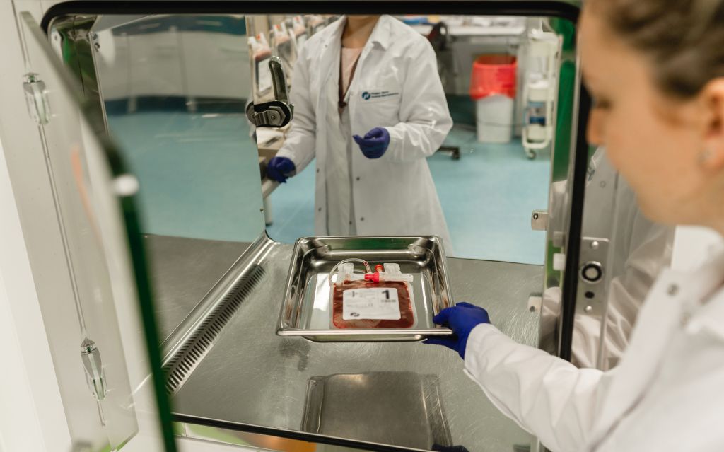 Dwie pracownice laboratorium w strojach medycznych przekazują sobie na tacy krew pępowinową w worku medycznym