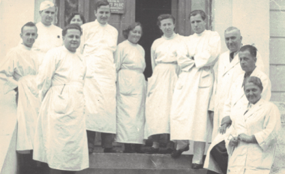 Zdjęcie historyczne, czarno-białe. 11 lekarzy w białych fartuchach na schodach przed wejsciem do budynku.