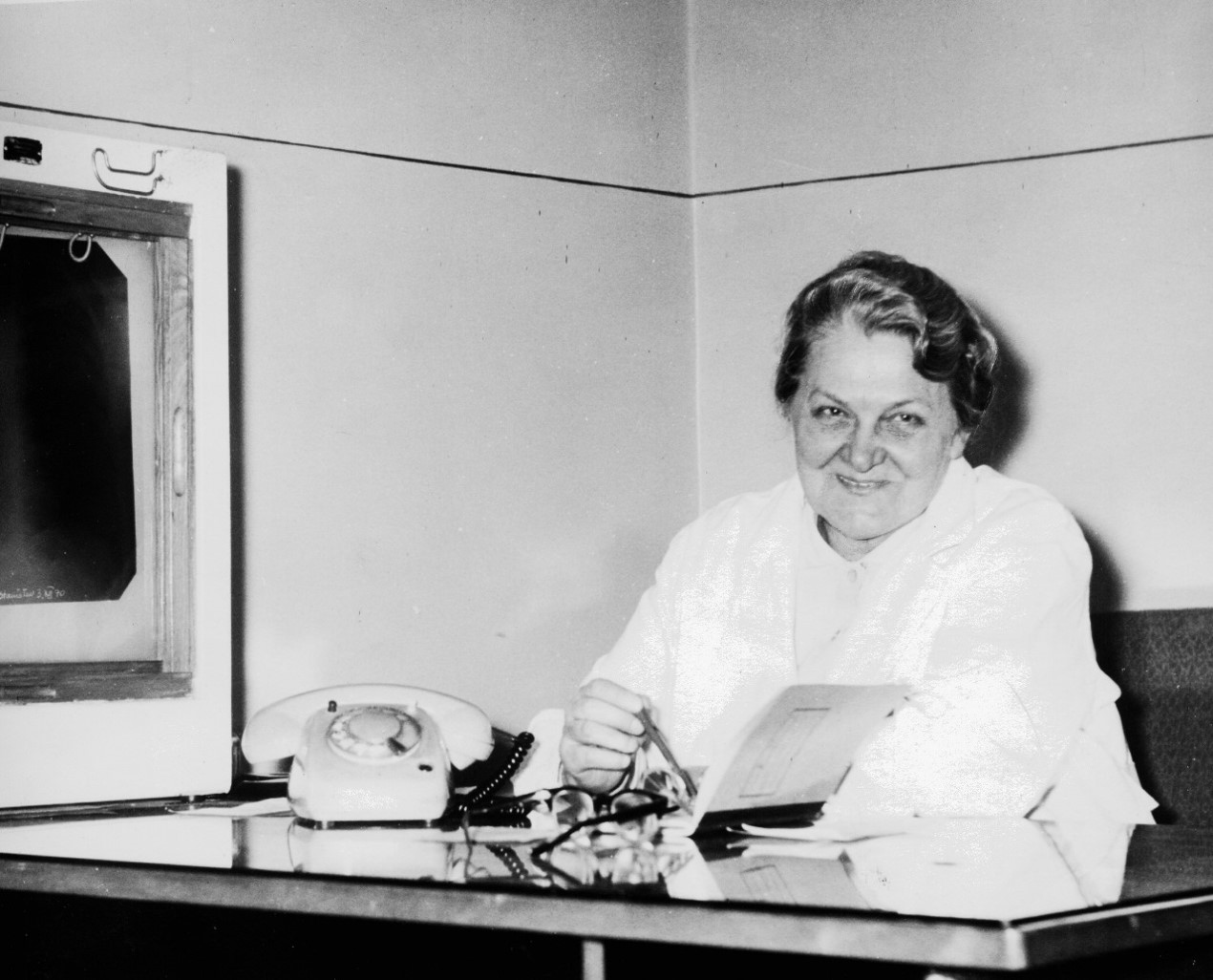 Zdjęcie historyczne, czarno-białe. Helena Mysakowska siedząca przy biurku z telefonem, uśmiecha się.