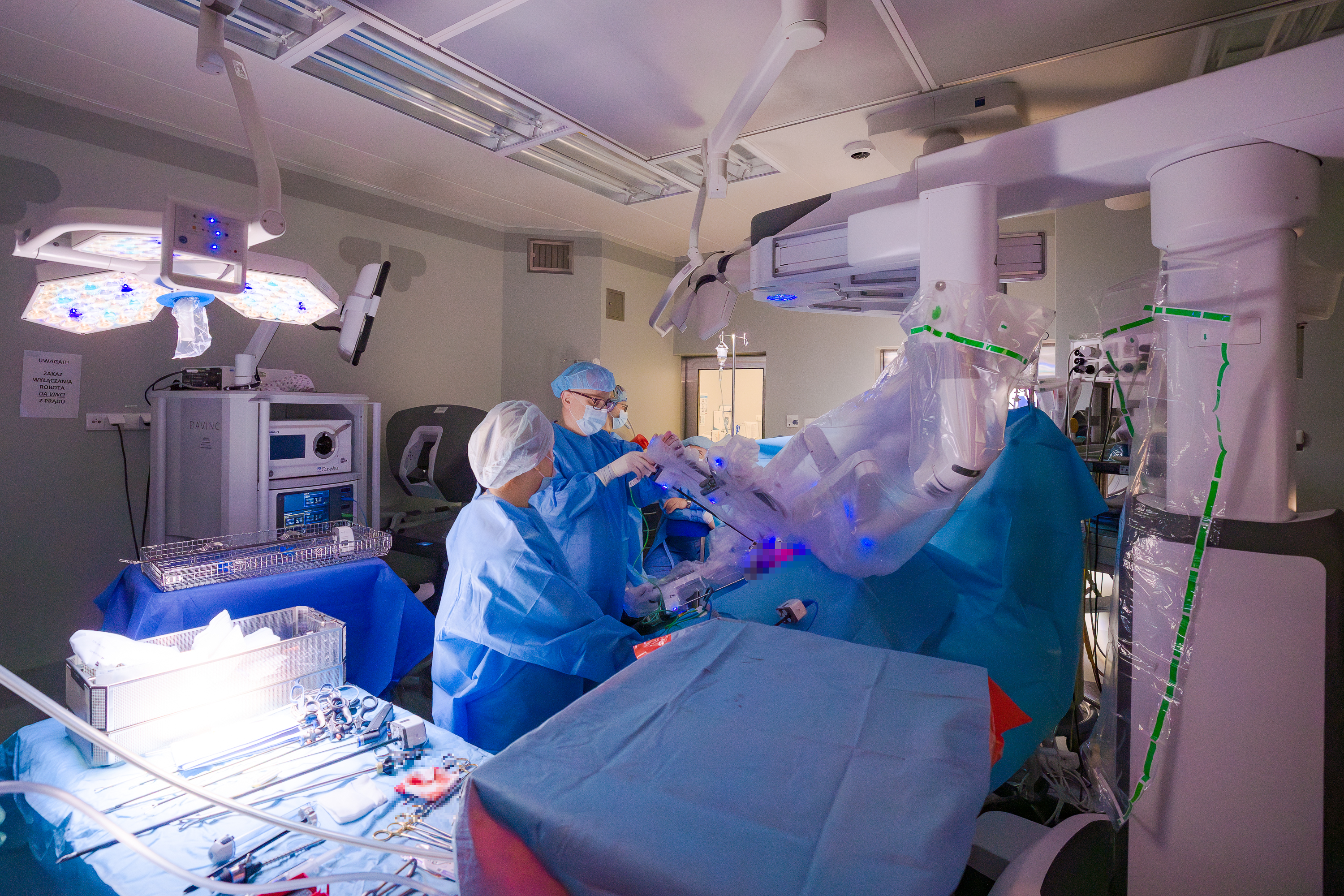 Sala operacyjna z robotem da Vinci. Po lewej stronie lekarz operujący wraz z pielęgniarką, pochyleni nad operowanym pacjentem. Obok, ułożone na stole narzędzia.