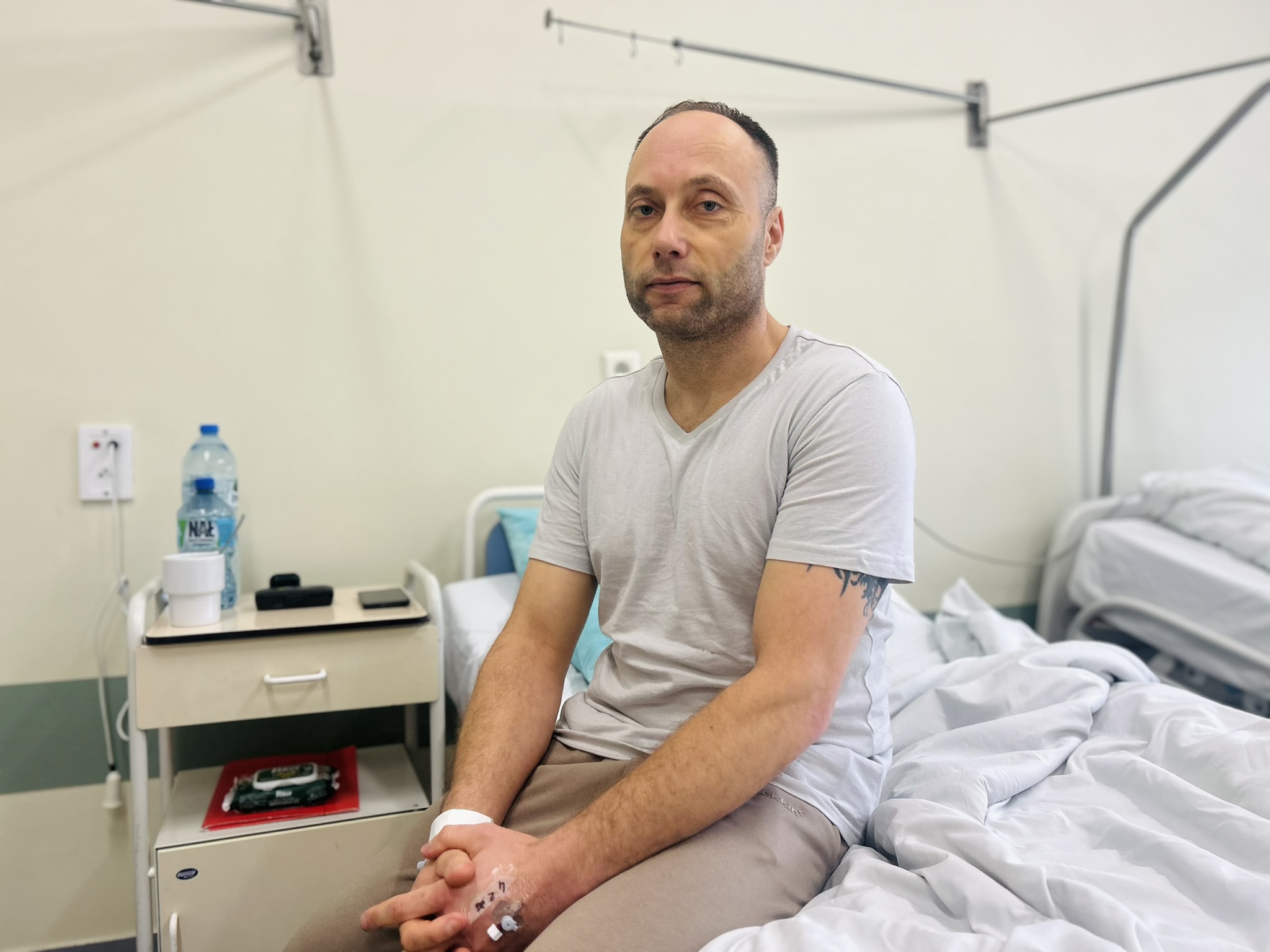 Na zdjęciu widać mężczyznę siedzącego na łóżku w jasnym pomieszczeniu. Ma na sobie szarą koszulkę i jasne spodnie, siedzi bokiem do łóżka i patrzy w stronę aparatu, trzymając dłonie splecione na kolanach. Na jego prawej ręce widoczny jest mały opatrunek, na prawej przezroczysty element przymocowany do skóry. Po lewej stronie fotografii stoi szafka z butelką wody, kubkiem i niewielkimi przedmiotami, a pod nią leży czerwony pakiet. Po prawej stronie fotografii znajduje się łóżko z jasną pościelą, lekko pogniecioną, a za mężczyzną widać ścianę z metalowymi elementami.