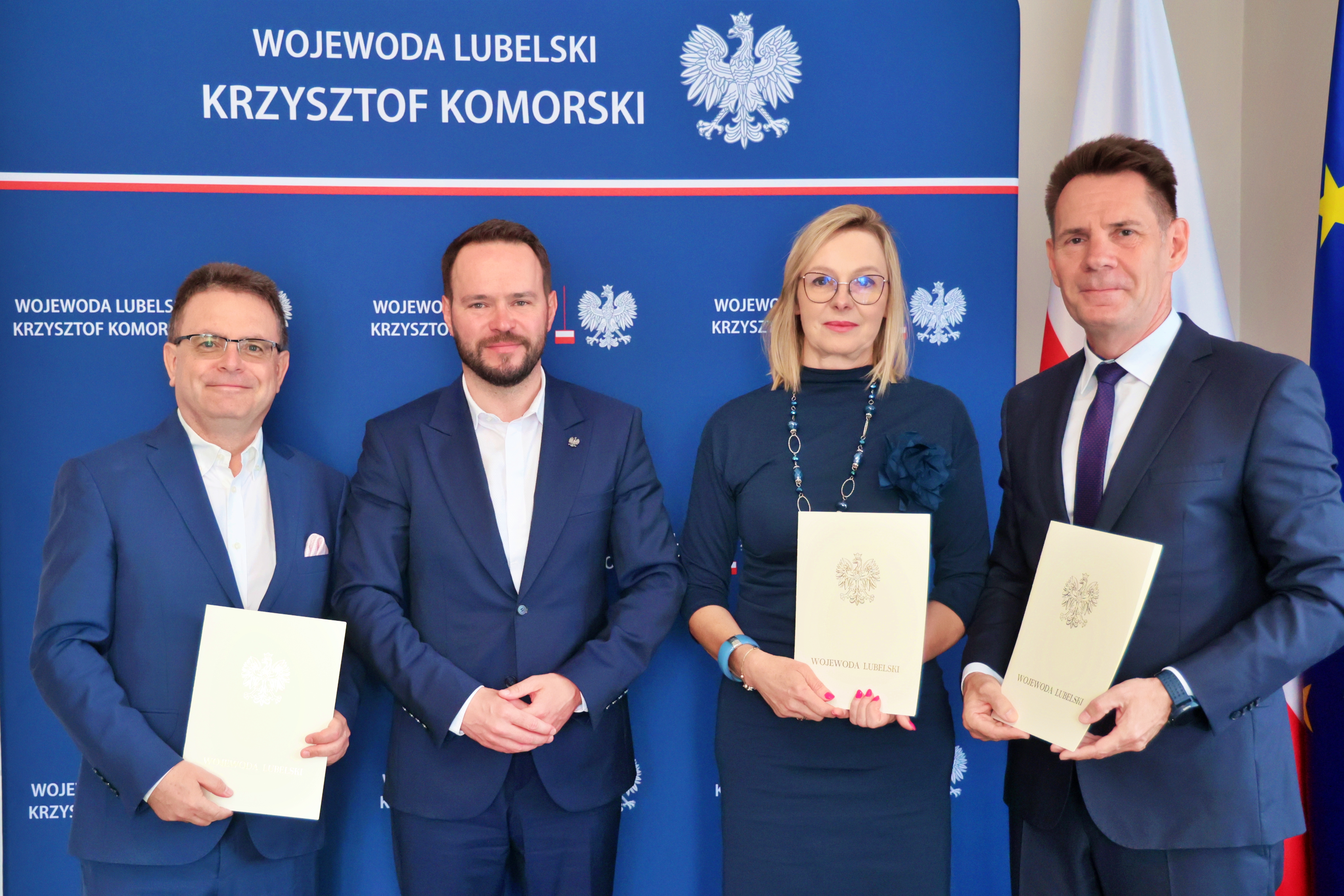 Cztery elegancko ubrane osoby - trzech mężczyzn i jedna kobieta. Trzy stoją trzymając w rękach teczki z orłem. Za nimi ścianka z napisem "wojewoda lubelski Krzysztof Komorski"