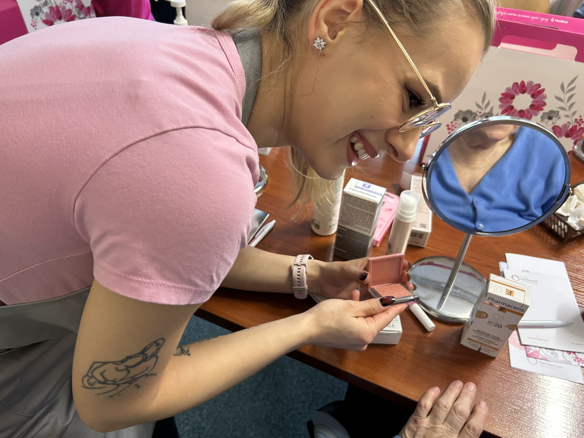 Na zdjęciu widać pochyloną do przodu młodą kobietę w różowej koszulce i szarym fartuchu, w okularach i z tatuażem na ramieniu. Uśmiecha się, patrząc w prawą stronę. W dłoniach trzyma pojemnik z różem. Za nią znajduje się stół, na którym widać kosmetyki i lustro, w którym odbija się szyja jakiejś osoby w niebieskiej koszulce. Z tyłu widać biało-różowe pudła.