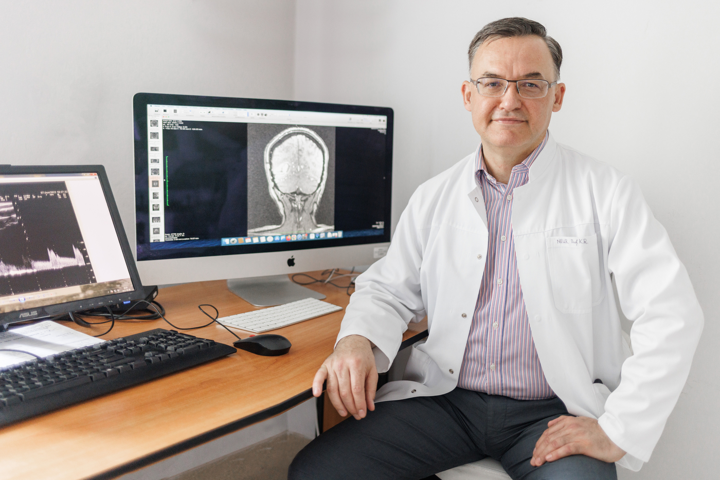 Na fotografii widać mężczyznę siedzącego na krześle przy biurku. Ma na sobie jasny fartuch i okulary. Jedna ręka spoczywa na kolanie, a druga na oparciu krzesła. Za nim stoi komputer, na którego ekranie widoczny jest czarno biały obraz przedstawiający głowę widzianą z boku. Z lewej strony widać drugi monitor z wykresami oraz klawiaturę i myszkę. Wokół znajdują się biurko i jasne ściany pomieszczenia.