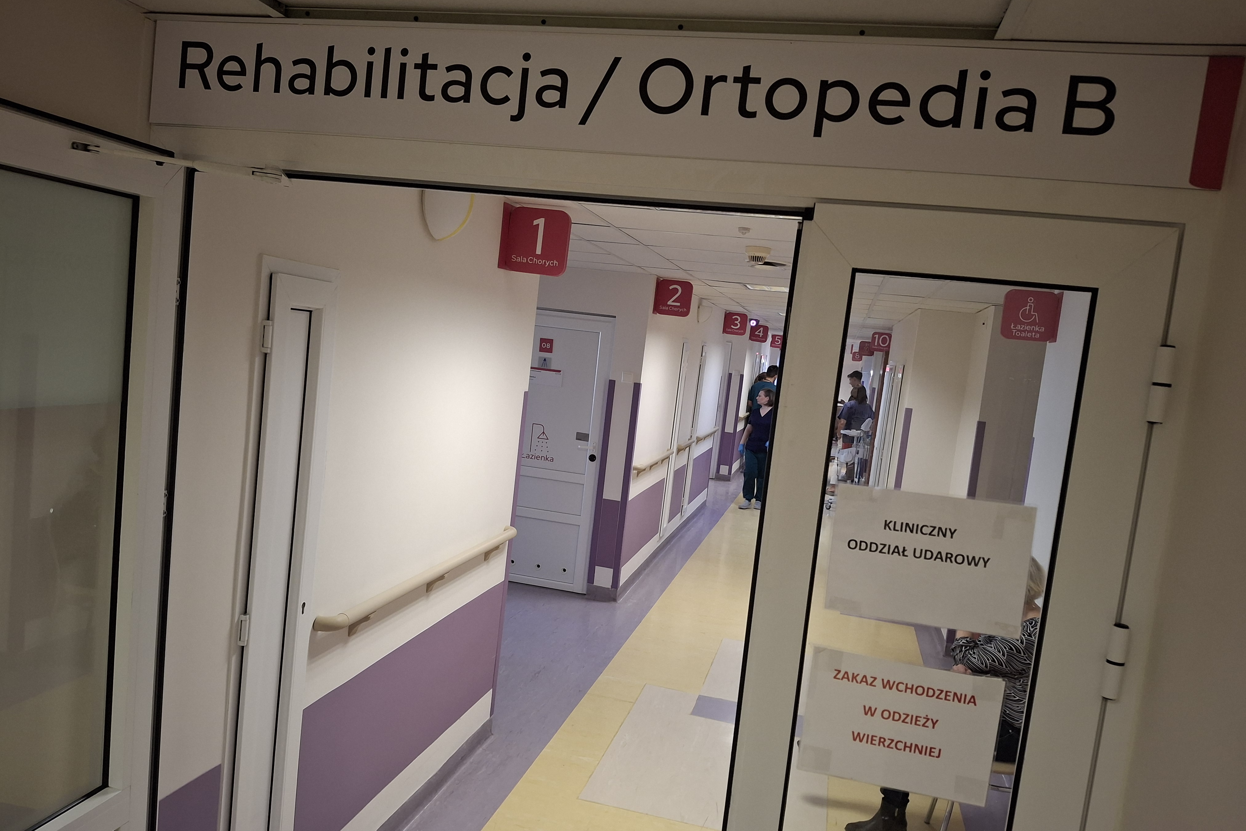 Wejście na Kliniczny Oddział Rehabilitacji, otwarte drzwi, w tle stojący na korytarzu lekarze. 